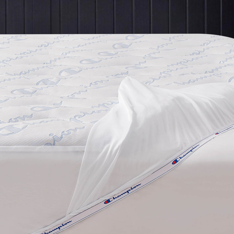 A close-up of a mattress with a white fitted sheet partially pulled back, revealing the Champion Cooling Knit Mattress Pad, which is stain-resistant and features the word “Champion” and the Champion logo printed repeatedly in blue.