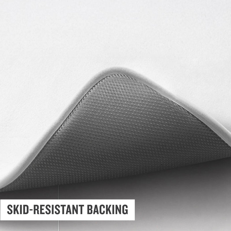 A close-up of a Logo Patch Memory Foam Bath Mat by Champion, with one corner lifted to reveal its textured, gray, skid-resistant backing. The words “SKID-RESISTANT BACKING” are overlaid in the bottom left corner.