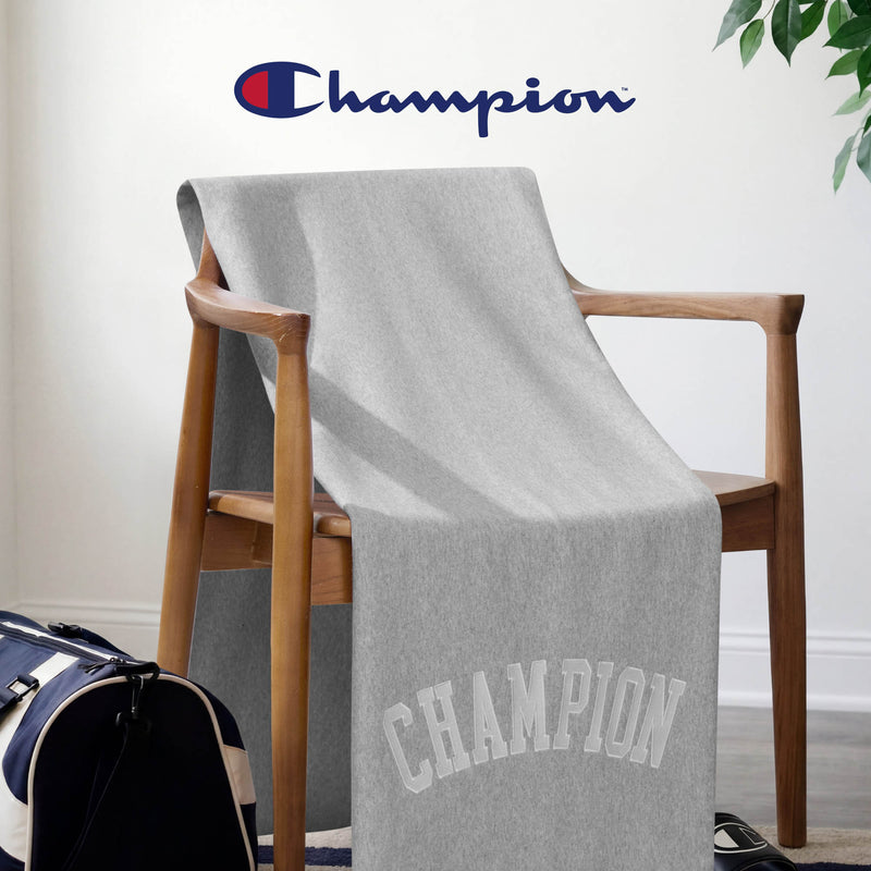 A Champion Jersey with Fleece Backing Throw Blanket in gray is draped over a wooden chair, while a blue and red Champion logo decorates the wall above. Nearby, a navy duffel bag sits on the floor, and a green plant in the corner adds to the cozy feel of this athletic bedding arrangement.