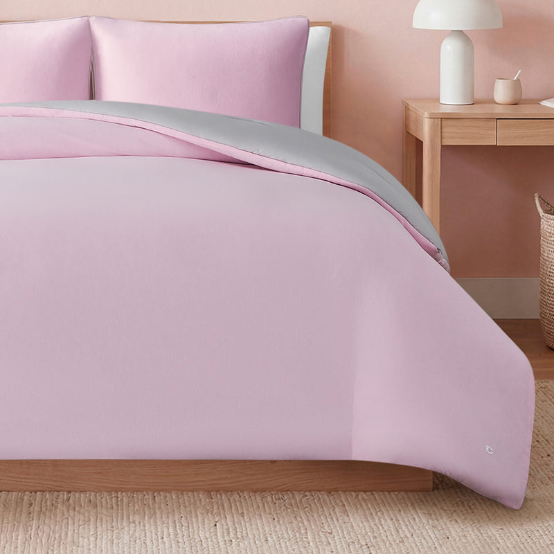 A neatly made bed features the Champion Reversible Solid Jersey Comforter Set in light pink with gray lining in a cozy bedroom, accompanied by a wooden nightstand holding a white lamp and cup, and a woven basket on the floor.
