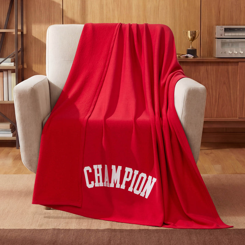 A Champion Jersey with Fleece Backing Throw Blanket is draped over a beige armchair in a cozy room with wood-paneled walls, a trophy, and a stereo on a shelf in the background.