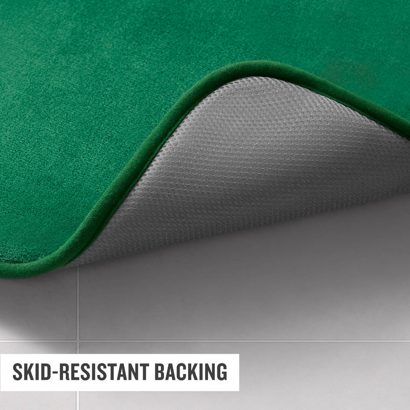 A green Champion Logo Patch Memory Foam Bath Mat is displayed on a tiled floor with one corner lifted to reveal its textured, skid-resistant gray backing. A label at the bottom reads SKID-RESISTANT BACKING.