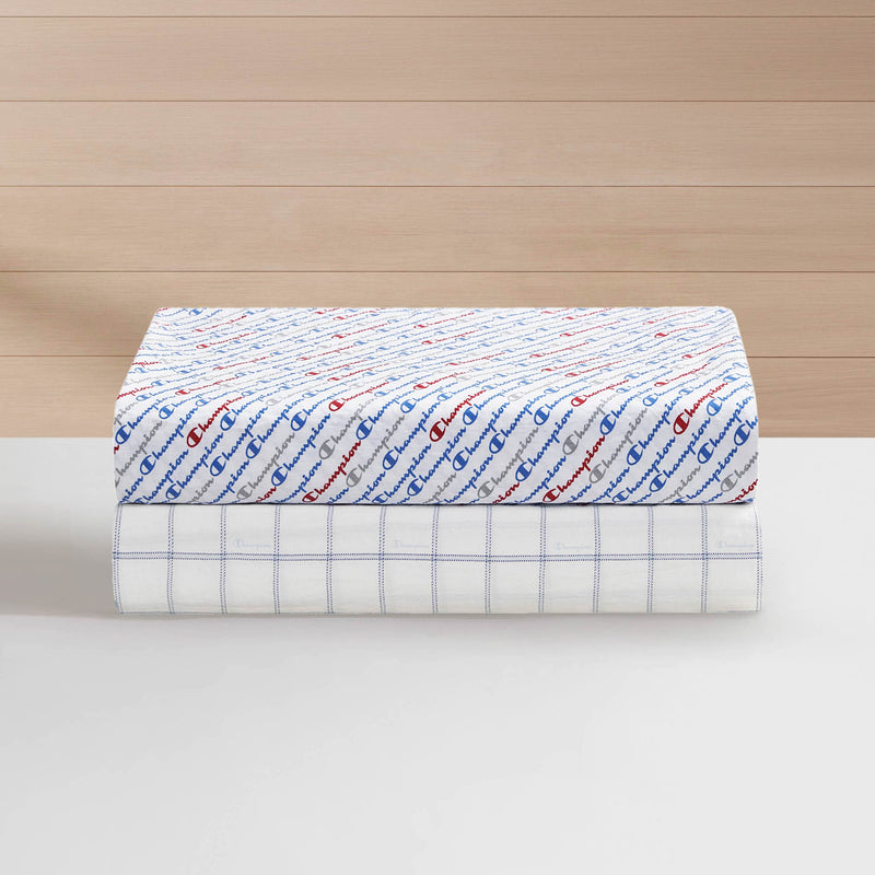 Two folded white sheets from the Champion Printed Microfiber Sheet Set rest stacked on a white surface; the top sheet displays a red and blue repeating Champion logo pattern, while the bottom sheet has a blue grid design. A light wood wall serves as the backdrop.