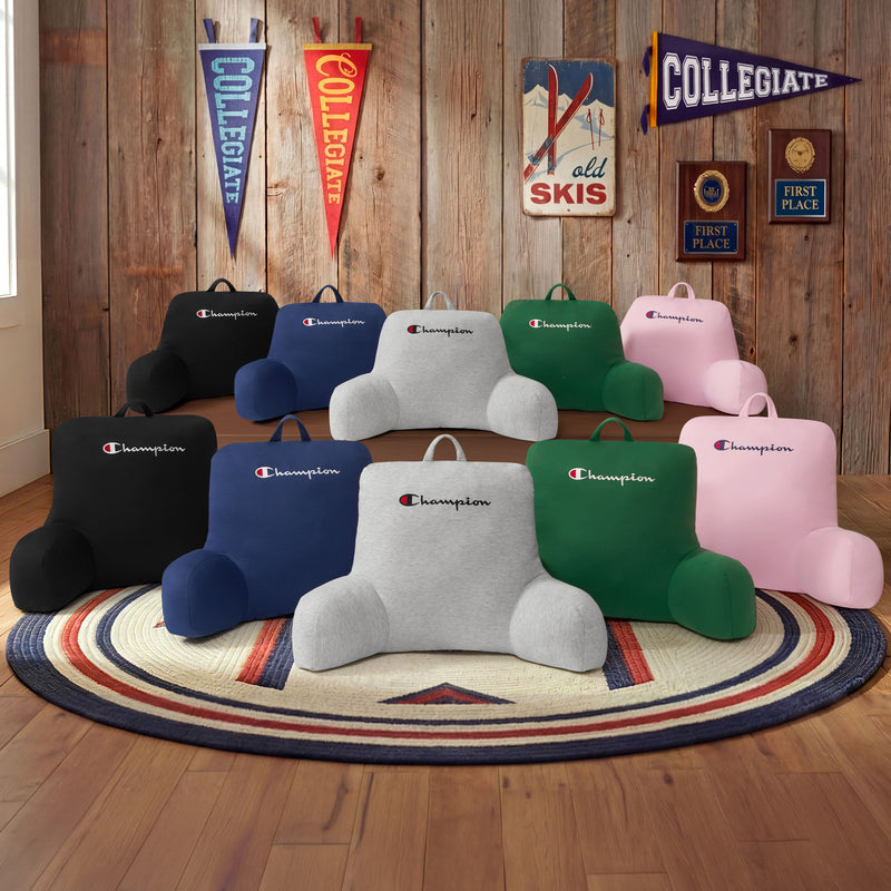 Nine Champion Jersey Backrest Pillows in black, blue, gray, pink, and navy—each adorned with the signature Champion logo—are arranged on a round rug in a wood-paneled room decorated with vintage decor, award plaques, and college pennants.