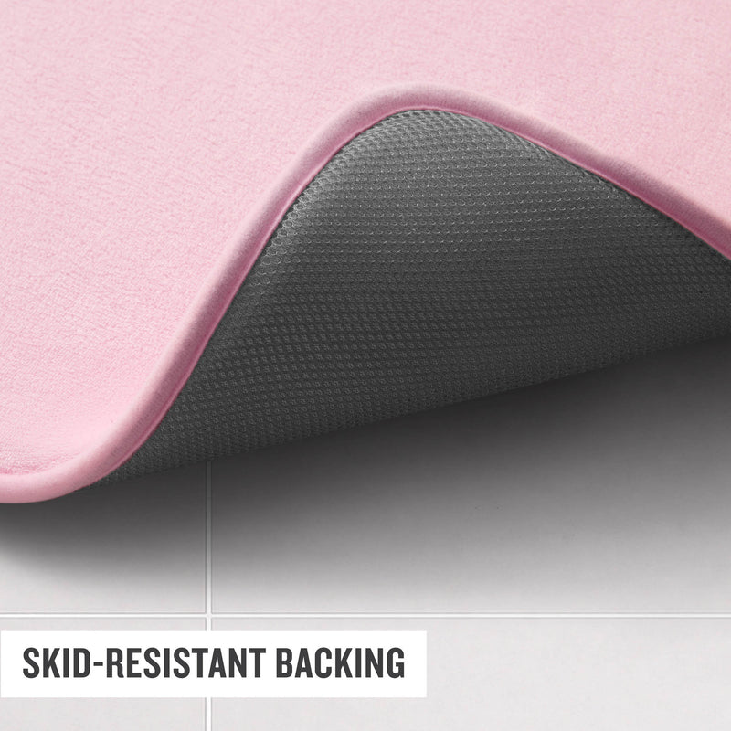 A close-up of a Champion Logo Patch Memory Foam Bath Mat in pink, with one corner lifted to display its textured gray skid-resistant backing, shown on a light-colored tiled floor. Text reads SKID-RESISTANT BACKING.