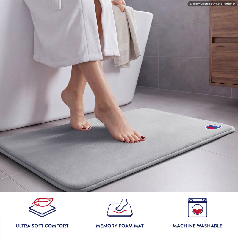 In a modern bathroom, a person in a white robe steps onto the gray Champion Logo Patch Memory Foam Bath Mat. This branded mat by Champion features ultra-soft comfort, supportive memory foam cushioning, and is machine washable, as highlighted by the icons below.