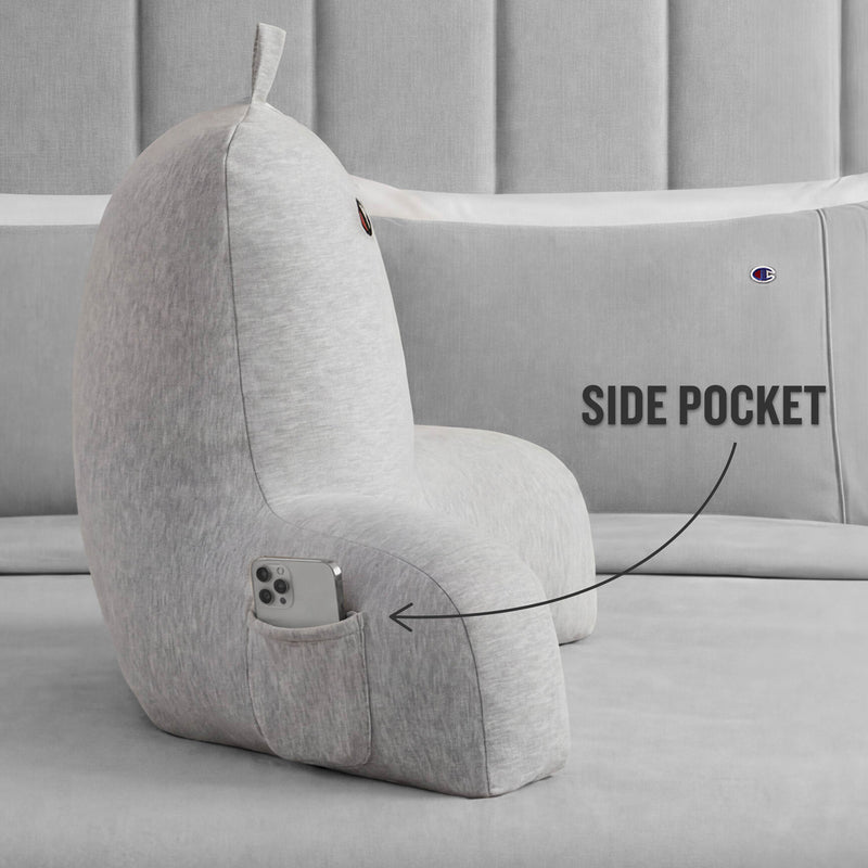A Champion Jersey Backrest Pillow in gray, featuring a convenient side pocket holding a smartphone, is placed on a gray bed. An arrow points to the pocket with the text SIDE POCKET written beside it.