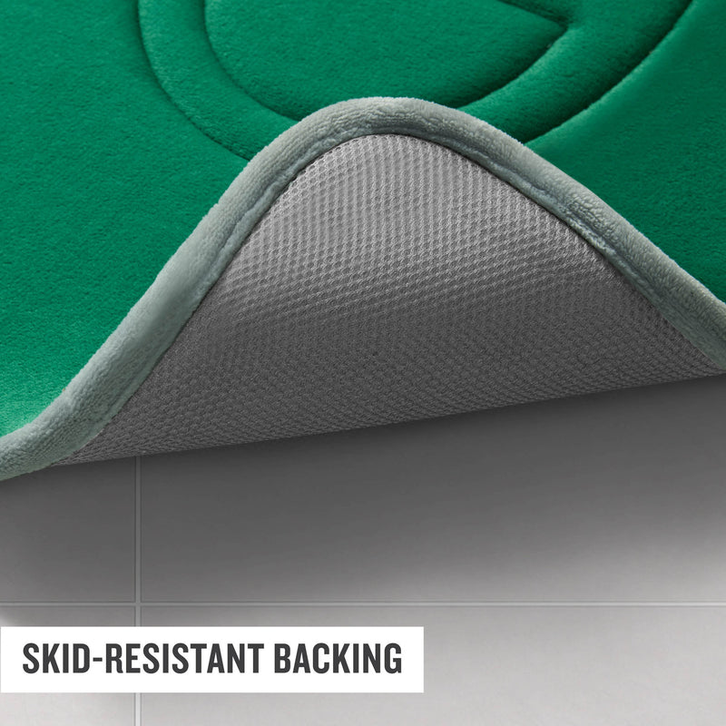 A close-up of the corner of a green Champion Quilted Logo Memory Foam Bath Mat, lifted to reveal its gray skid-resistant backing, shown on a light-colored tiled floor. Designed for any bathroom, this mat delivers plush comfort and dependable traction underfoot. Text in the image reads SKID-RESISTANT BACKING.