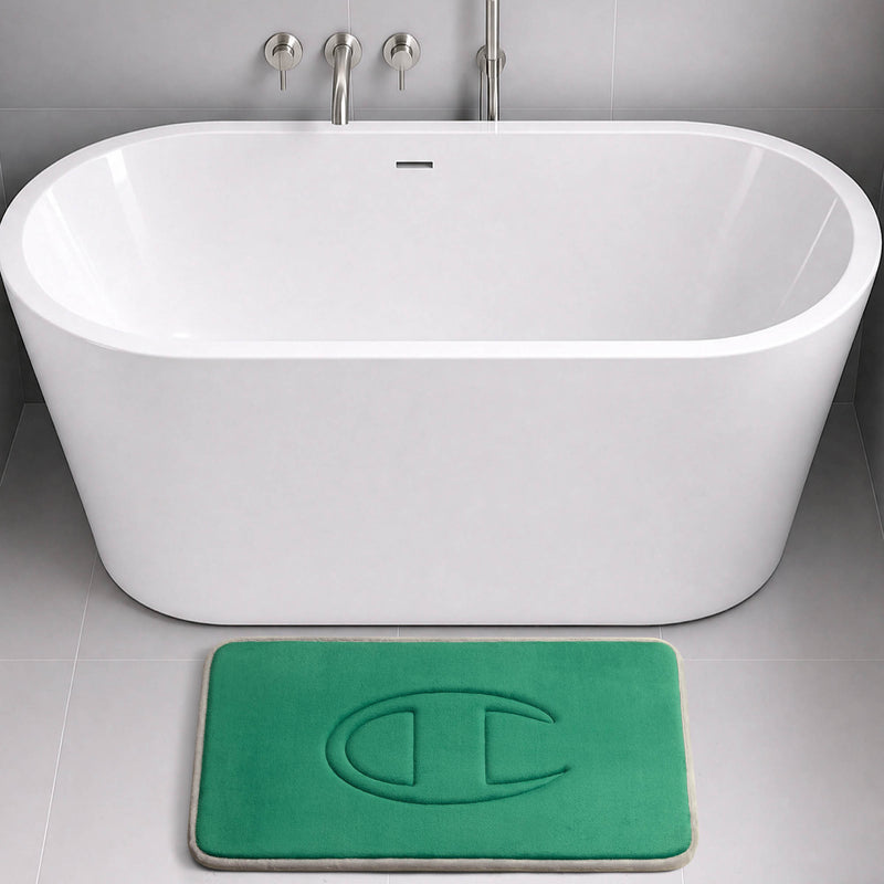 A modern white bathtub with wall-mounted fixtures sits above a gray tile floor. In front of the tub is a green Champion Quilted Logo Memory Foam Bath Mat, featuring an embossed C logo design.