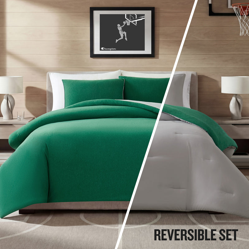 A modern bedroom featuring a bed dressed in the Champion Reversible Solid Jersey Comforter Set in green and white. The space includes two nightstands, matching lamps, and framed basketball artwork. Text reads: REVERSIBLE SET.