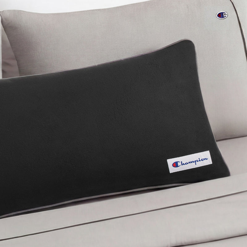 A Champion Faux Wool Body Pillow in black is placed on a light gray couch, displaying the Champion logo on both the pillow and the couch. Filled with plush polyester, this body pillow offers maximum comfort and style to any space.