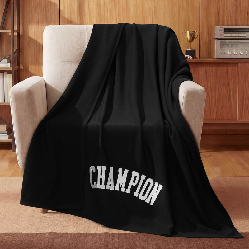 A Champion Jersey with Fleece Backing Throw Blanket in black, featuring the word CHAMPION in bold white letters, is draped over a beige armchair in a wood-paneled room, with a trophy and stereo displayed on a shelf in the background.