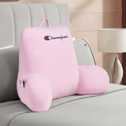 A Champion Jersey Backrest Pillow in pink, featuring supportive armrests and a soft jersey knit cover, sits on a neatly made bed with a bedside table and lamp in the background.