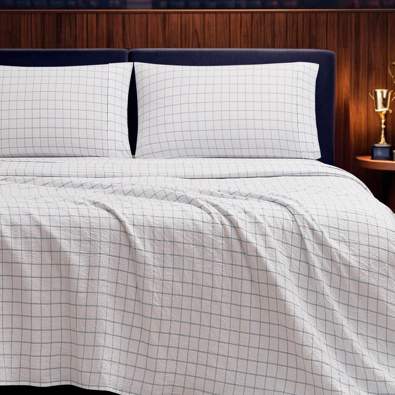 A neatly made bed is styled with the Champion Printed Microfiber Sheet Set in a white, wrinkle-resistant fabric featuring a blue grid pattern, paired with two matching pillows. The setup is complemented by a dark headboard, a wooden wall, and a small nightstand displaying a gold trophy.