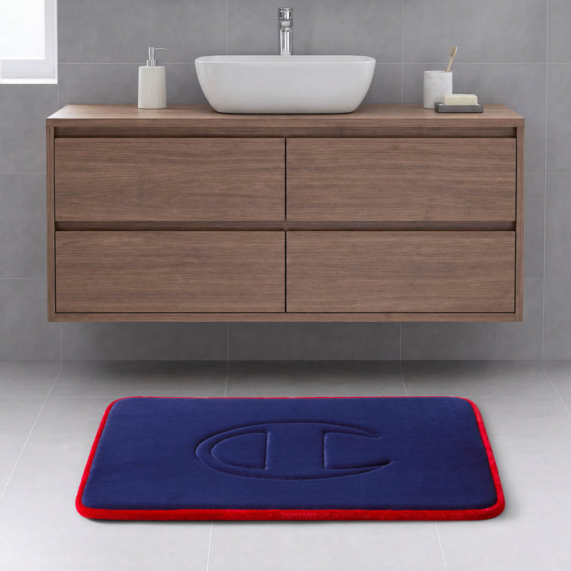 A modern bathroom with a wooden vanity, a white vessel sink, and the Champion Quilted Logo Memory Foam Bath Mat, which features a blue surface with a red border and embossed design, placed on a light gray tile floor.