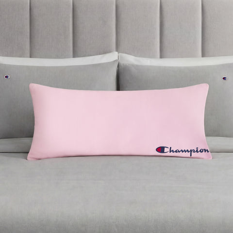 A Champion Jersey Body Pillow in pink is centered on a neatly made bed with gray bedding and matching pillows, each pillowcase adorned with small Champion logos. This cozy accent from Champion adds sporty flair to the room, making it perfect for fans of the brand.