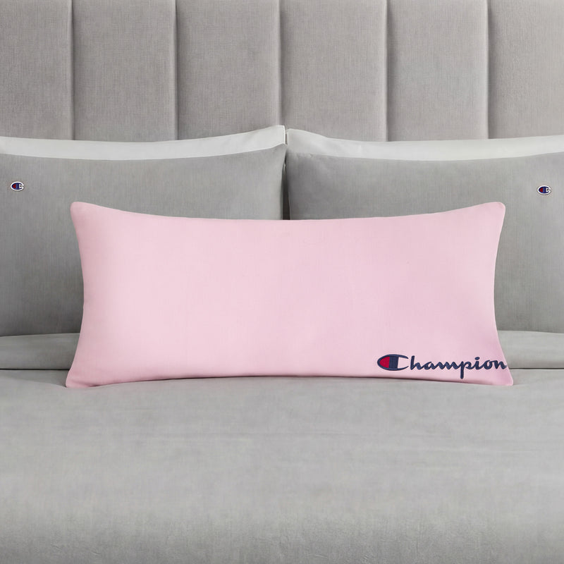 A Champion Jersey Body Pillow in pink is centered on a neatly made bed with gray bedding and matching pillows, each pillowcase adorned with small Champion logos. This cozy accent from Champion adds sporty flair to the room, making it perfect for fans of the brand.