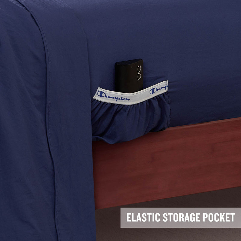 A navy blue Peak Comfort Microfiber Sheet Set by Champion features a white and blue elastic storage pocket on the side, designed to conveniently hold a black item. Ideal for athletic bedding needs, these deep pocket sheets combine style with functionality. The bottom right corner is labeled with the text “ELASTIC STORAGE POCKET.”.