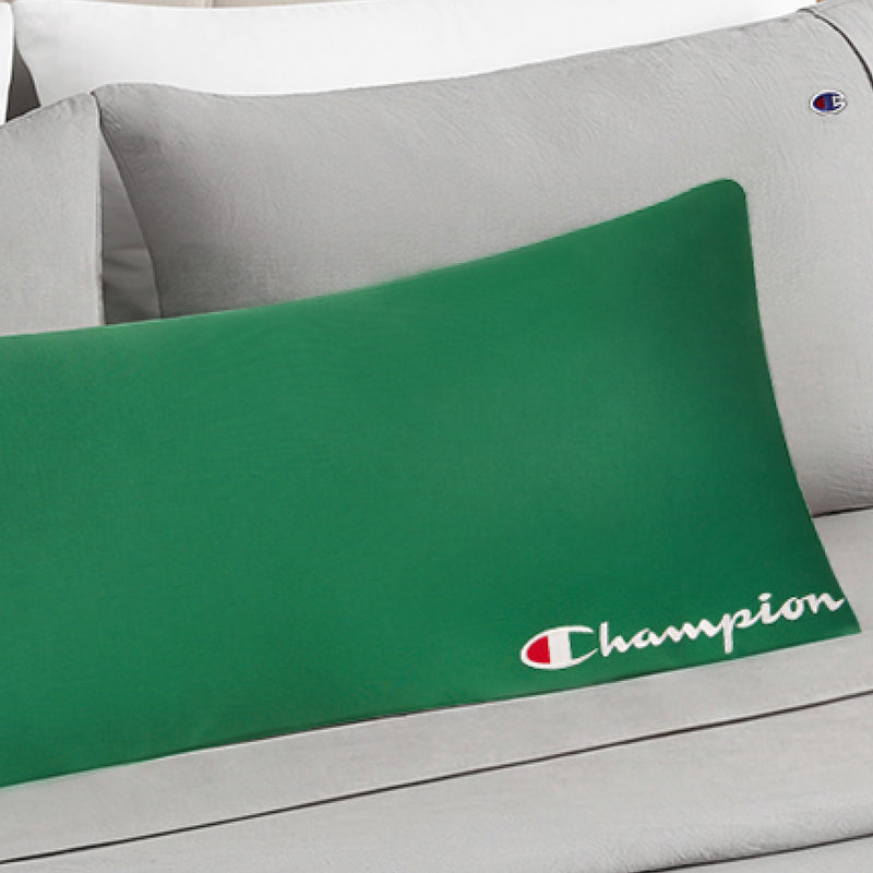 A Champion Jersey Body Pillow in green, showcasing the iconic white Champion logo and text, rests on a bed dressed in light gray bedding adorned with subtle Champion logos, adding an athletic touch to the room.