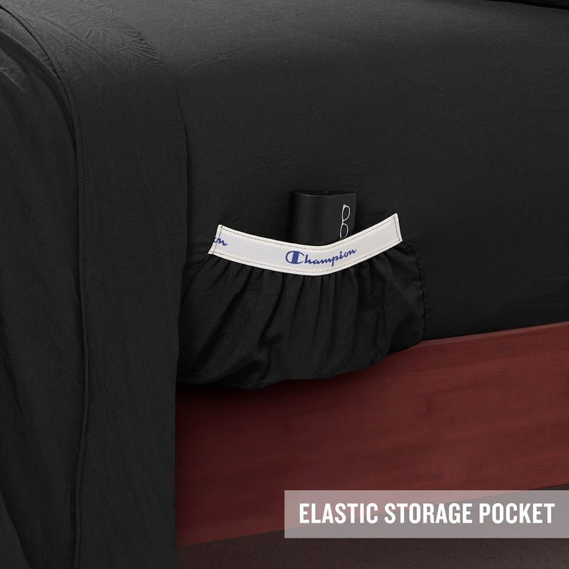 A close-up of a bed with black Champion Peak Comfort Microfiber Sheet Set featuring an elastic storage pocket holding a glasses case. The pocket, designed as part of these athletic bedding essentials, has a white trim with Champion written on it. Text in the corner reads ELASTIC STORAGE POCKET.