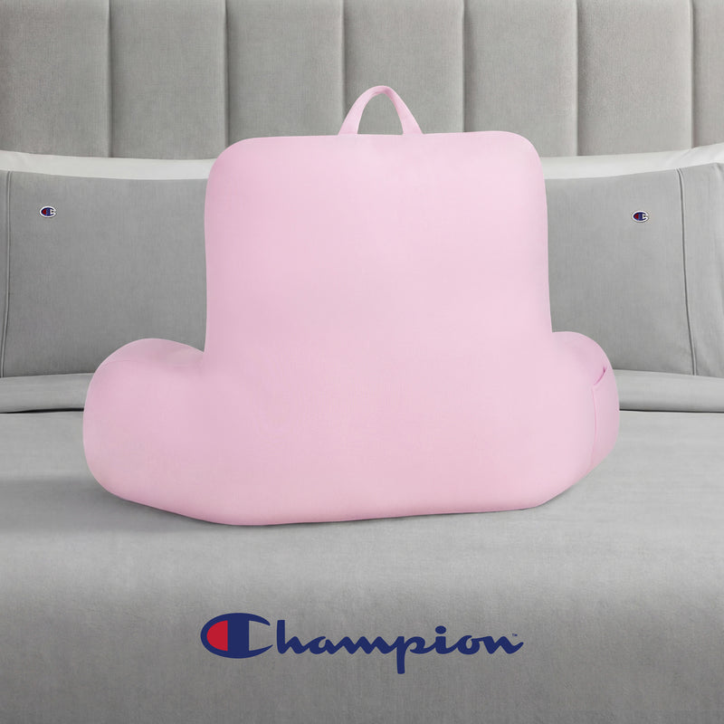 A pink Champion Jersey Backrest Pillow with armrests sits on a gray bed dressed in soft jersey knit bedding, featuring Champion logos on the pillows and headboard, with a large Champion logo displayed at the foot of the bed.