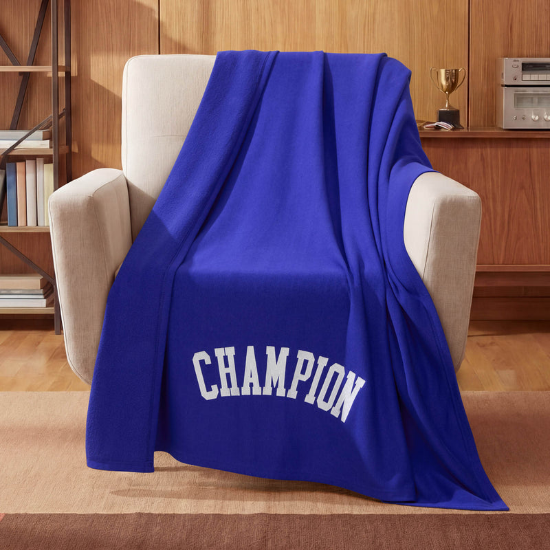 A Jersey with Fleece Backing Throw Blanket by Champion in royal blue, featuring the word CHAMPION in white letters, is draped over a beige armchair in a cozy wood-paneled room, with a trophy displayed on a shelf in the background.