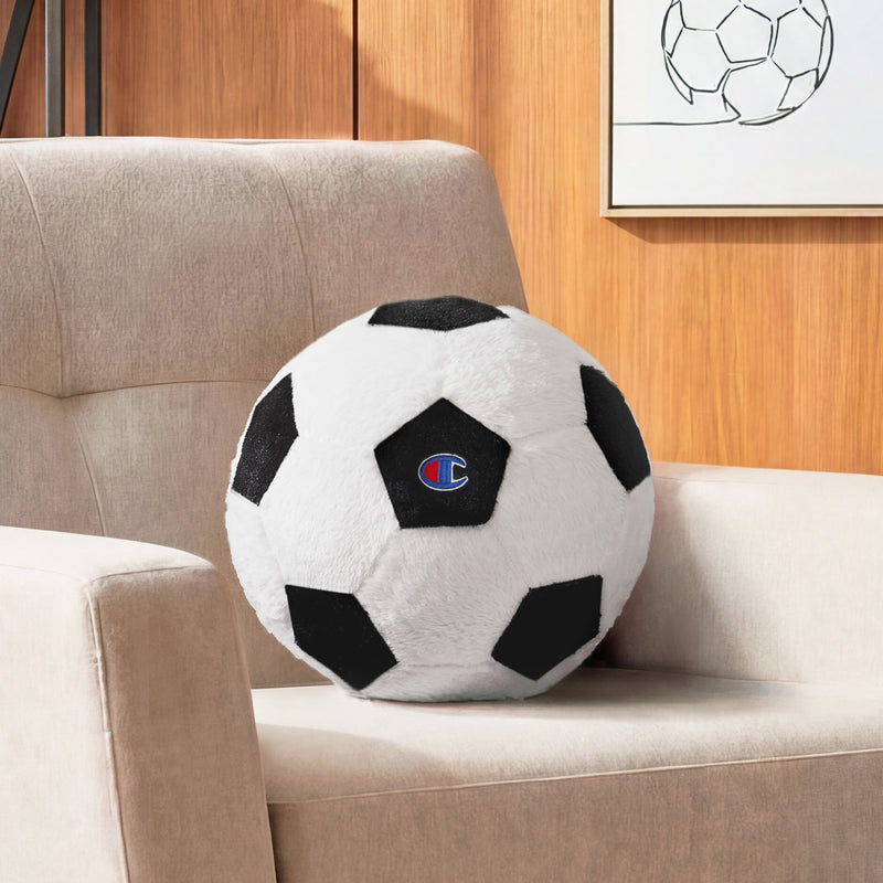 A Champion Sport Shaped Pillow featuring classic black and white patches rests on a beige armchair, while a framed soccer ball illustration adds sporty charm to the wooden wall in the background.
