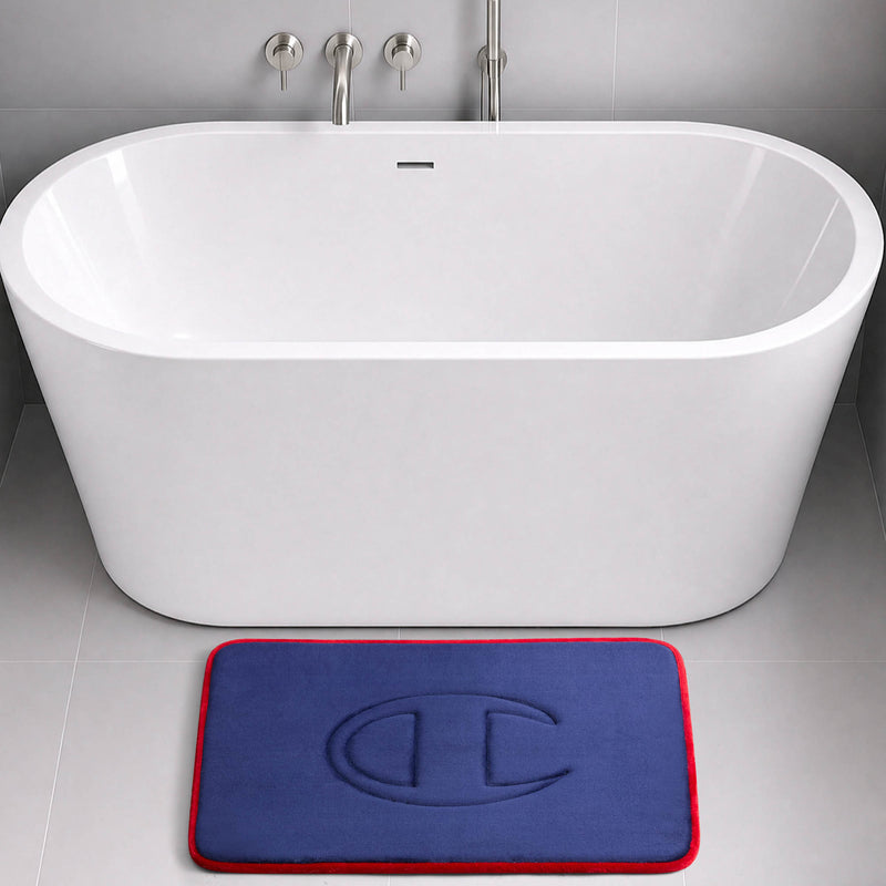 A modern white bathtub with wall-mounted faucets sits on a gray tiled floor. In front of the tub is a blue Champion Quilted Logo Memory Foam Bath Mat featuring a red border and an embossed design.