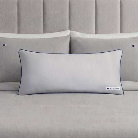 A Champion Faux Wool Body Pillow in light gray, featuring plush polyester fill, dark piping, and a small Champion logo, rests in front of two matching gray pillows on a neatly made bed with a gray upholstered headboard.