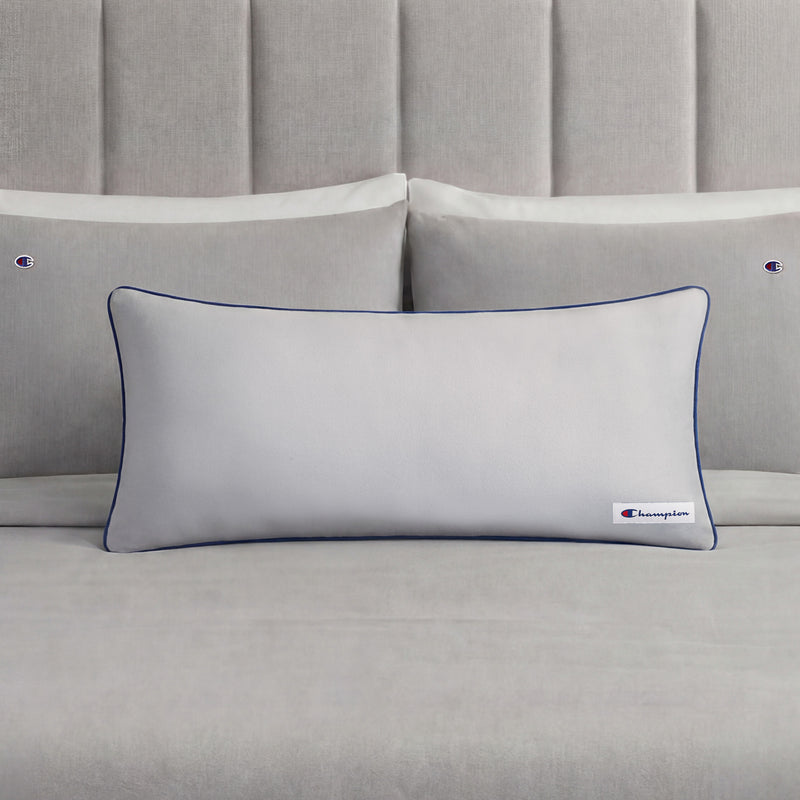 A Champion Faux Wool Body Pillow in light gray, featuring plush polyester fill, dark piping, and a small Champion logo, rests in front of two matching gray pillows on a neatly made bed with a gray upholstered headboard.