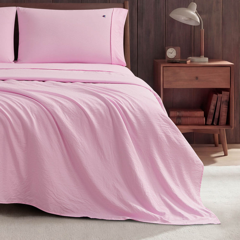 A neatly made bed with light pink Peak Comfort Microfiber Sheet Set by Champion sits beside a wooden nightstand holding a lamp, clock, and books. The scene has a cozy, minimalist look with a wooden accent wall in the background.