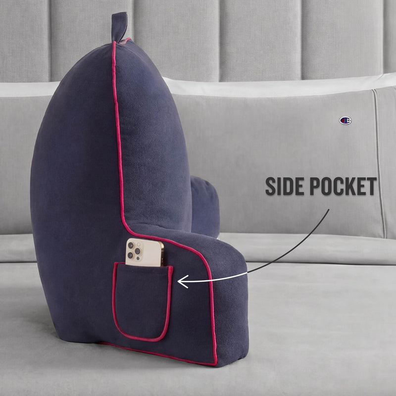A Faux Wool Backrest Pillow by Champion in navy blue with red trim sits on a bed. The pillow features a convenient side pocket, which is shown holding a smartphone. An arrow points to the pocket with the label SIDE POCKET.