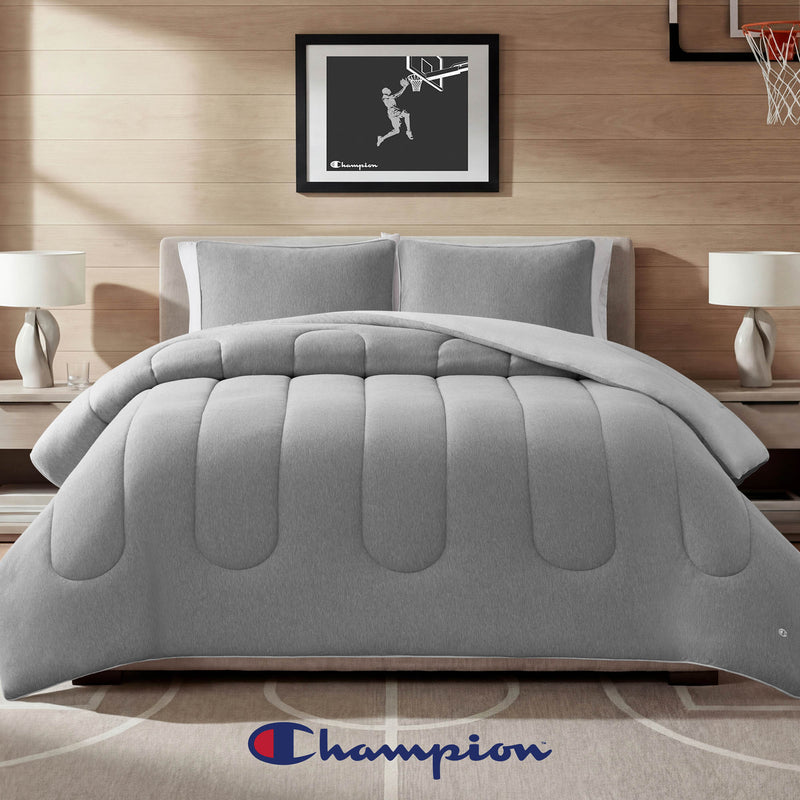 A modern bedroom showcasing a gray Premium Reversible Solid Jersey Comforter Set by Champion, paired with two lamps on nightstands and basketball-themed wall art, features an athletic bedding vibe accentuated by the iconic Champion logo at the foot of the bed.