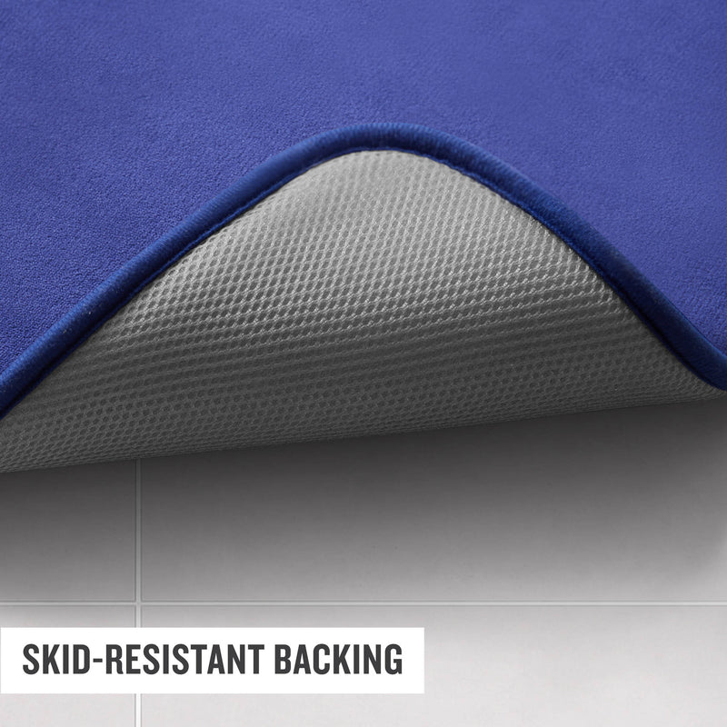 A close-up of a Champion Logo Patch Memory Foam Bath Mat on a tiled floor, with one corner lifted to reveal its gray, textured skid-resistant backing. Text reads: SKID-RESISTANT BACKING.