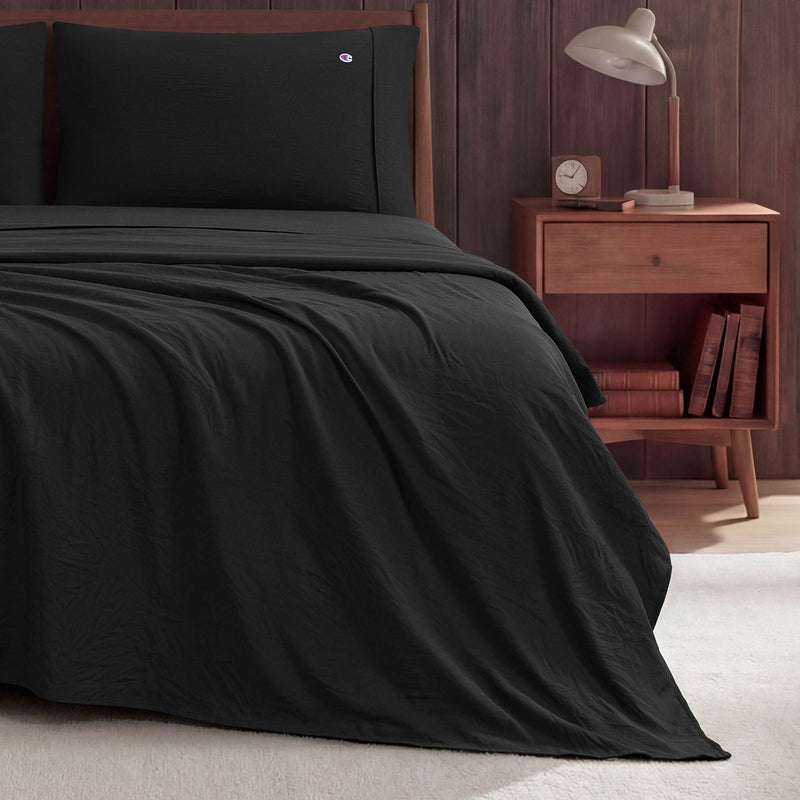 A neatly made bed with black Champion Peak Comfort Microfiber Sheet Set is next to a wooden nightstand holding a lamp, clock, and two books. The background features a wood-paneled wall and beige carpeted floor.