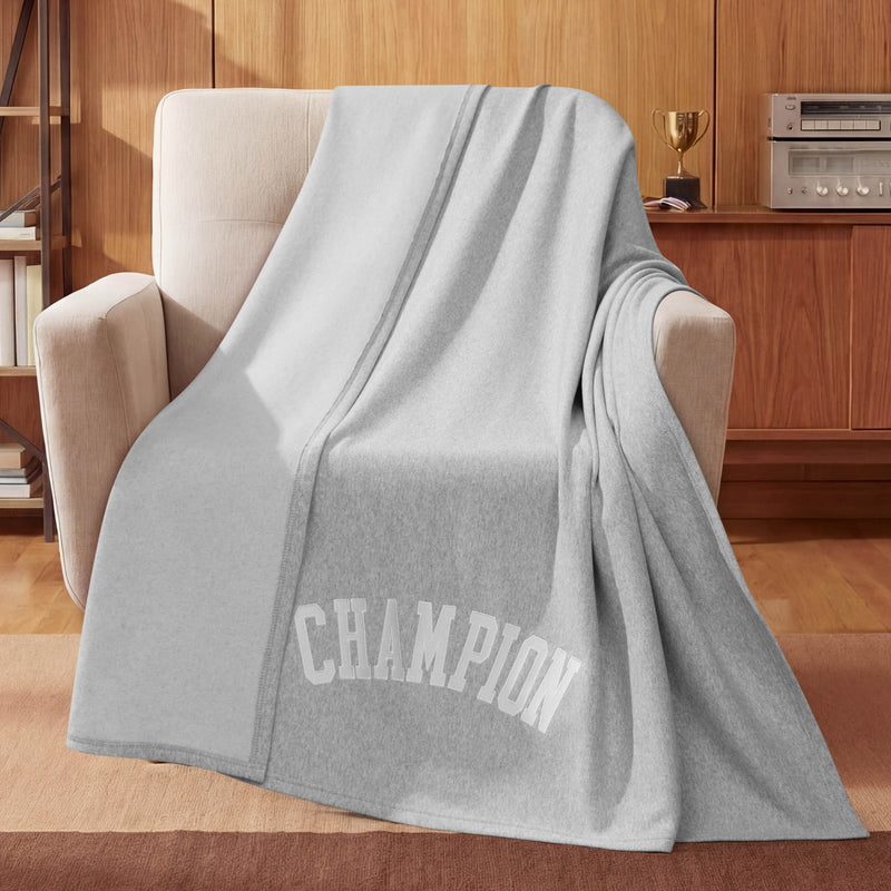 A cozy room with wooden furniture, a trophy, and a stereo system features a beige armchair draped with the Champion Jersey with Fleece Backing Throw Blanket in light gray, boldly displaying the word CHAMPION—an ideal choice for those looking for stylish and comfortable athletic bedding.