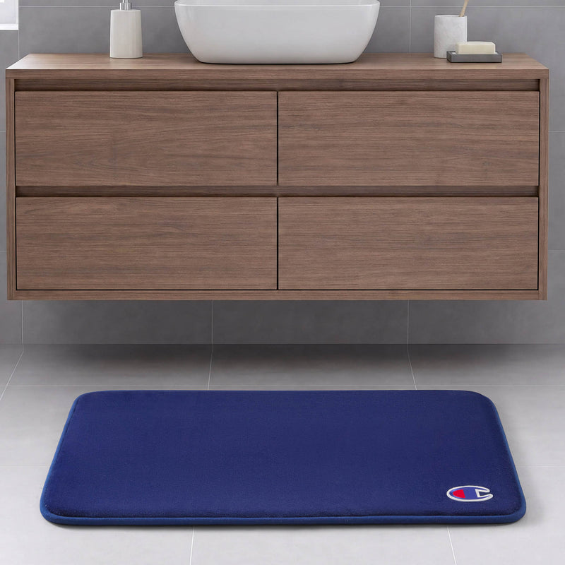 A blue Logo Patch Memory Foam Bath Mat by Champion, featuring a small logo in the corner, is placed on a light gray tile floor in front of a modern wooden bathroom vanity.